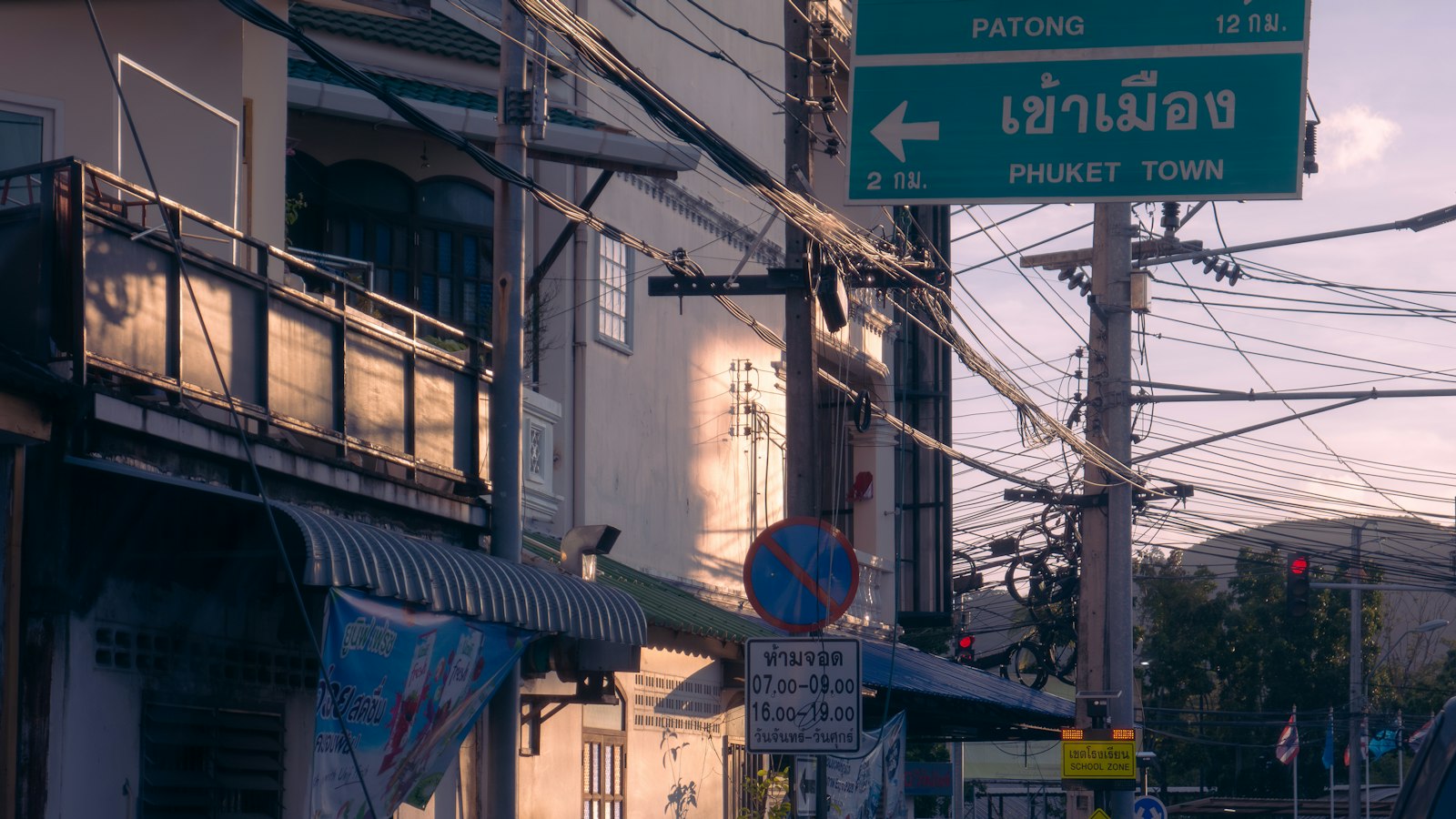 ถนนในภูเก็ตที่มีป้ายจราจรสีเขียวเขียนว่า เข้าเมือง, Phuket Town อยู่เหนือตึกแถวและสายไฟที่พันกันยุ่ง