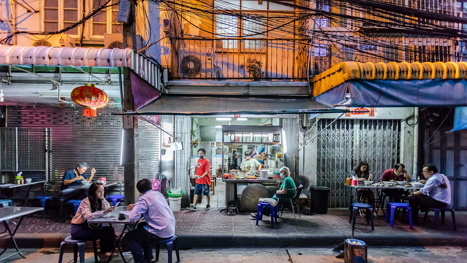 ผู้คนนั่งกินข้าวและคุยกันที่โต๊ะพลาสติกหน้าร้านอาหารริมทางในกรุงเทพฯ ตอนกลางคืน